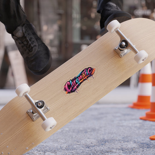 A skateboard flips midair, revealing a bold, graffiti‑style “Chromatic” graphic splashed with vivid pink, blue, orange, and black. The light wood deck and clean white wheels contrast with the energetic artwork, while a skater in black shoes adds motion and attitude. Traffic cones and a textured gray surface create an urban backdrop, emphasizing the dynamic, colorful, artistic style ideal for expressive fashion branding.