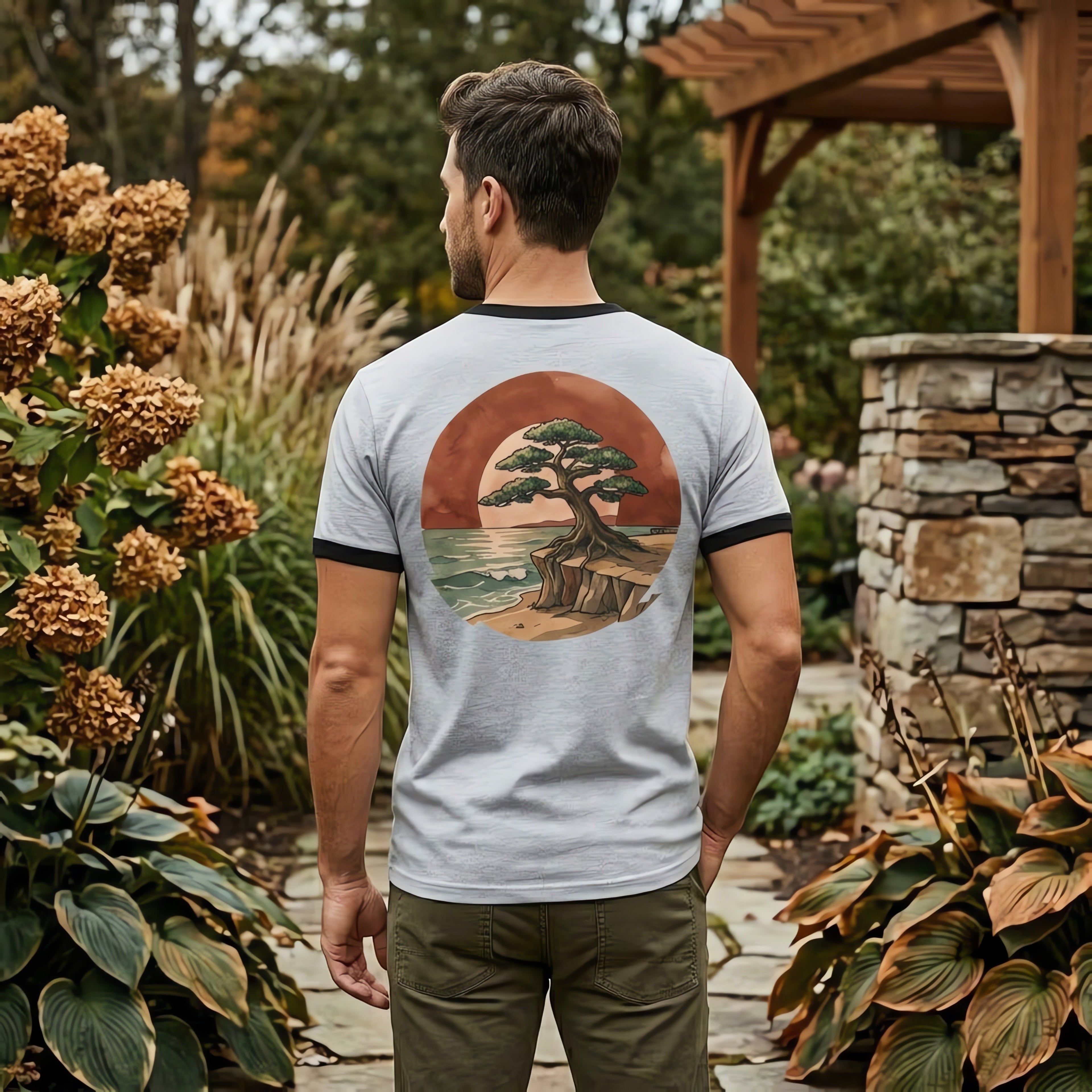 Man wearing a Chromatic light grey ringer tee with a back graphic of a bonsai tree on a cliffside at sunset.