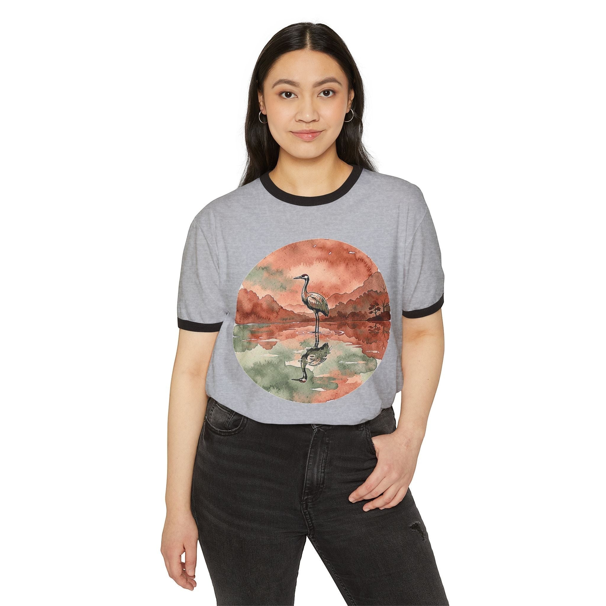 A woman wearing a grey Chromatic ringer tee with a watercolor crane and pond graphic, featuring black trim.